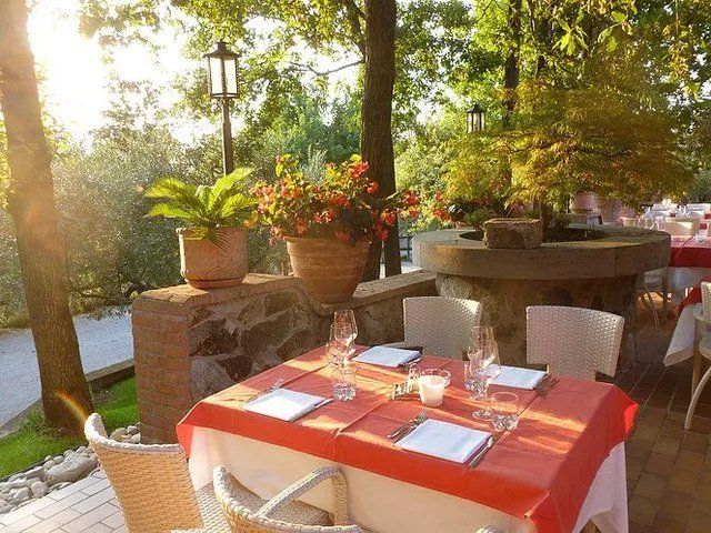 Tavolo da ristorante all'aperto apparecchiato con tovaglia rossa, circondato dal verde.