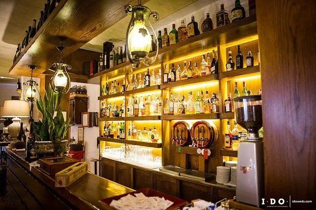 Interno del bar con scaffali illuminati che ospitano bottiglie di liquore. Bancone in legno e macinacaffè visibili.