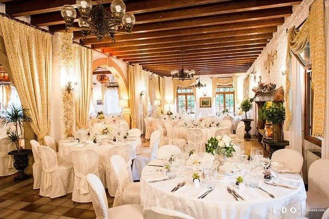 Elegante sala per banchetti, tavoli rotondi con tovaglie bianche, fiori, lampadario decorato, travi in legno.