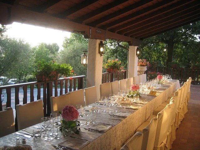 Lungo tavolo apparecchiato per un pasto in veranda, con fiori, posate e sedie.