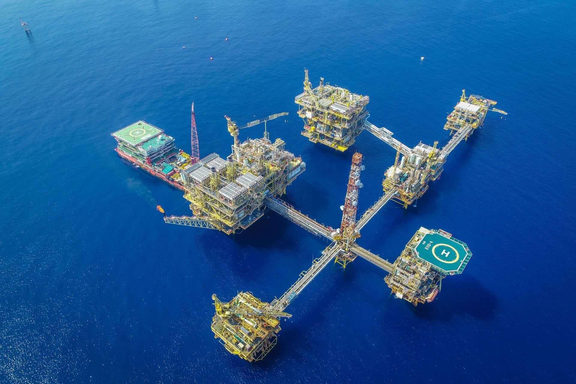 Vue aérienne de plusieurs plateformes pétrolières offshore interconnectées en eau bleue, avec un héliport.