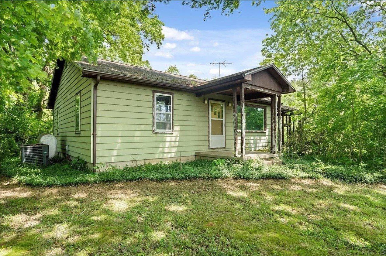 A small green house with a porch is surrounded by trees.