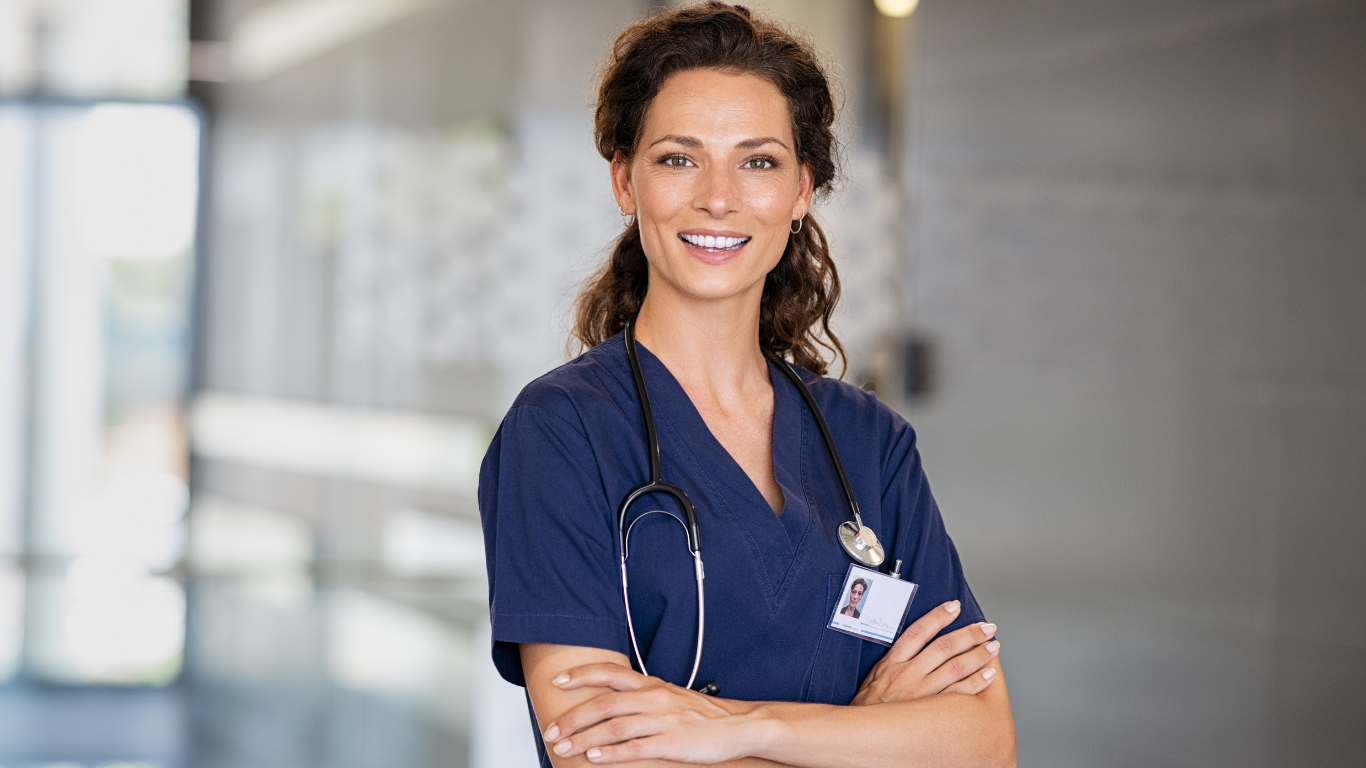 A nurse is standing in a hospital hallway with her arms crossed and smiling.