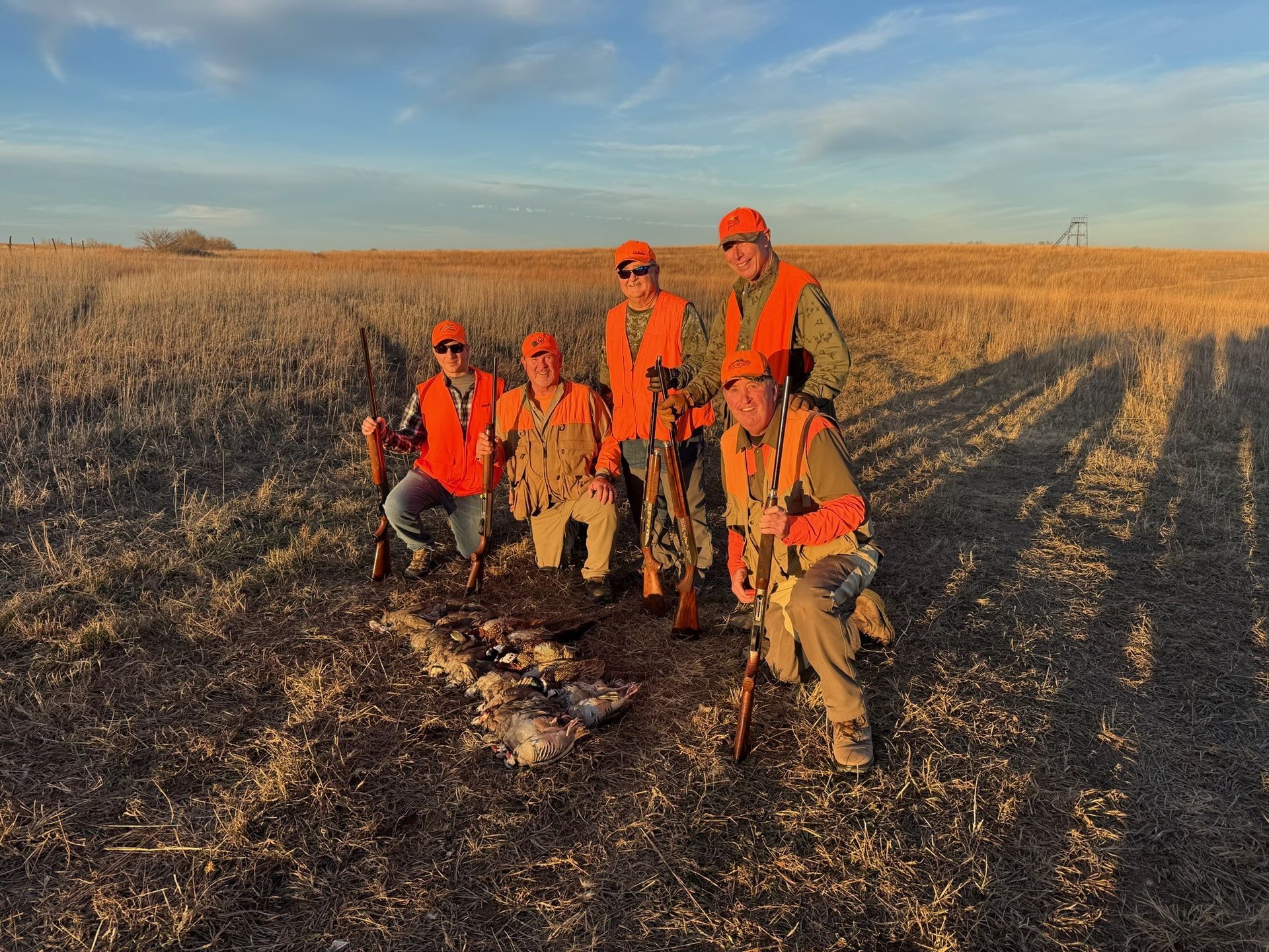 Five people in orange vests pose with hunting rifles and dead birds in a field.