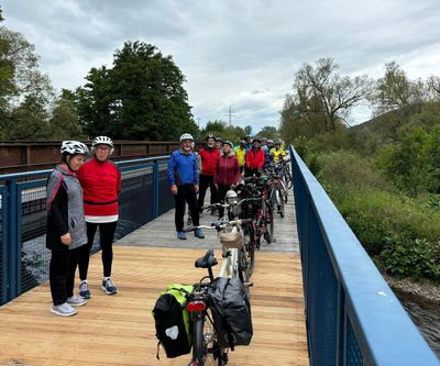Gruppe von Tandem-Radfahrern in wetterfester Kleidung auf einer Brücke, umgeben von Bäumen und bewölktem Himmel.