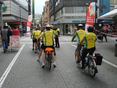 Gruppe von Tandem-Radfahrern in gelb-grünen Trikots fährt durch eine Fußgängerzone, mit Helmen und Gepäcktaschen.