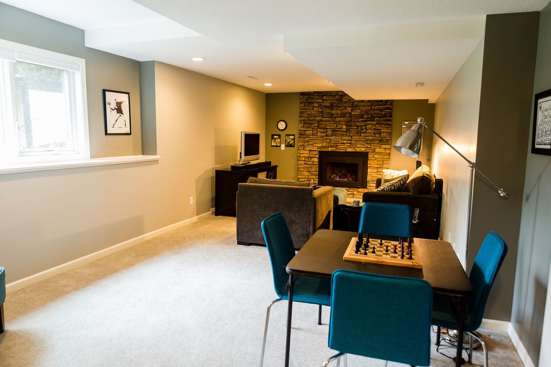 Basement living area with a fireplace, seating, a table, and a window; neutral tones with teal accents.