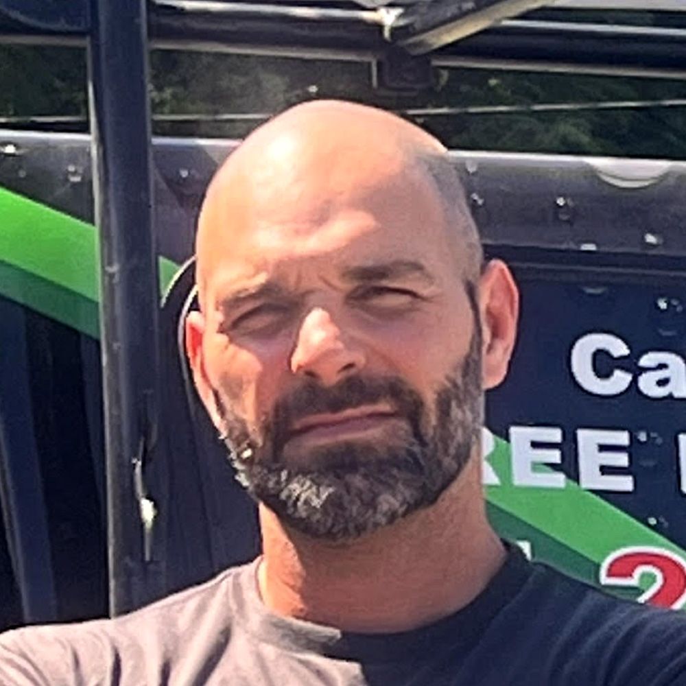 A bald man with a beard is standing in front of a vehicle.