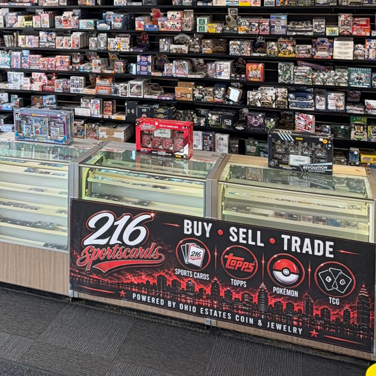 Trading card store counter with shelves of boxed cards and a large “BUY SELL TRADE” sign for 216 Sports Cards.