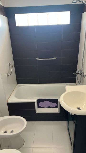 Baño pequeño con bañera, lavabo, inodoro y una pared de ducha revestida de azulejos oscuros debajo de una ventana.