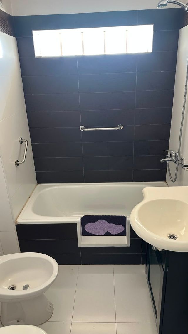 Baño pequeño con bañera, lavabo, inodoro y una pared de ducha revestida de azulejos oscuros debajo de una ventana.
