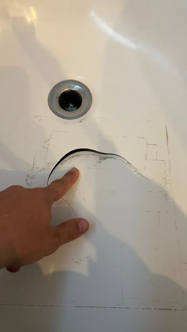Una mano señala una grieta alrededor del desagüe de un lavabo de baño en una superficie de porcelana blanca.
