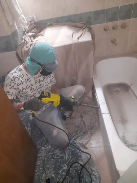 Un trabajador con capucha y mascarilla de color turquesa rocía limpiador en espuma en un baño junto a una bañera.