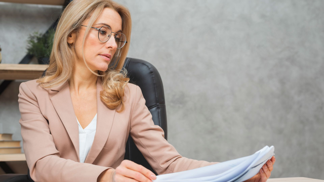 Woman in glasses and tan blazer examines papers at a desk, office setting.