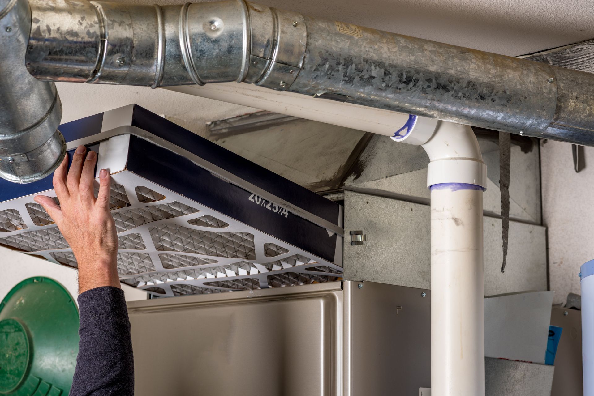Hand checking a black HVAC filter above a metal duct and white PVC pipe in a utility room
