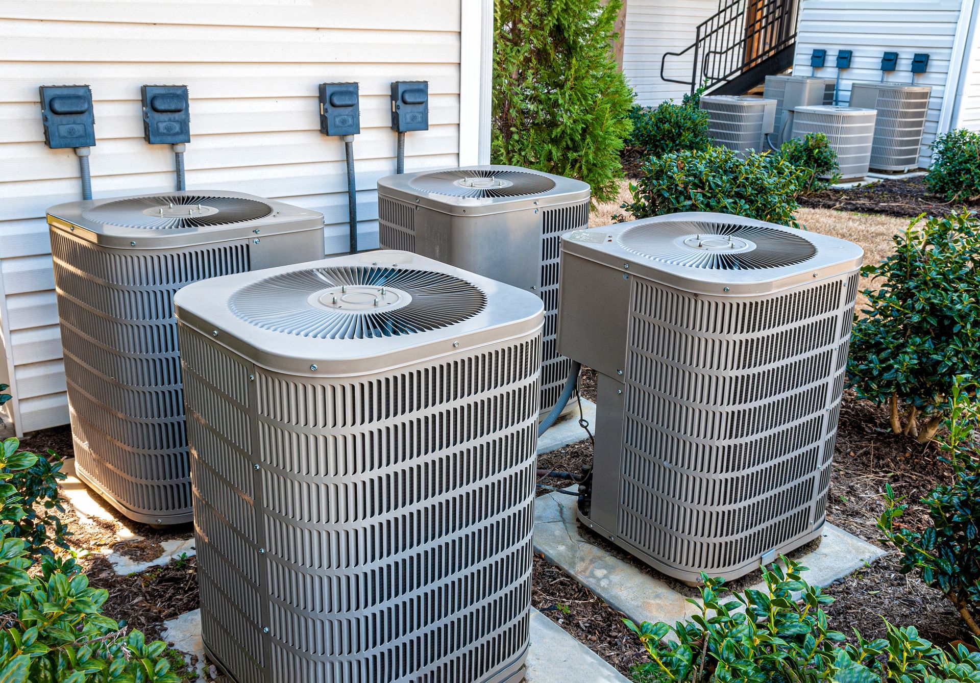 Four outdoor air conditioning units beside a house with siding and shrubs