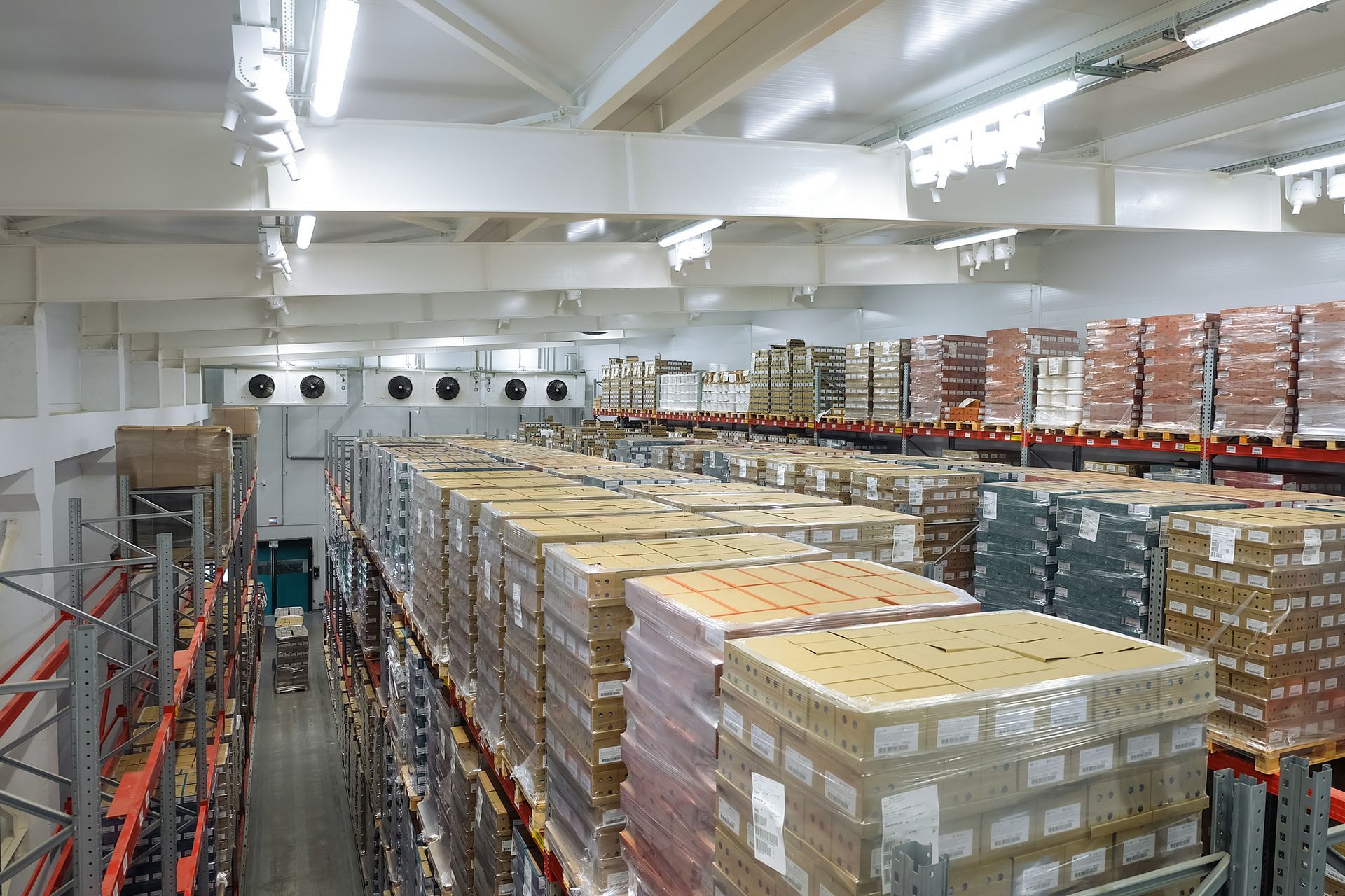 Warehouse interior with stacked pallets of boxed goods and shelving under bright ceiling lights