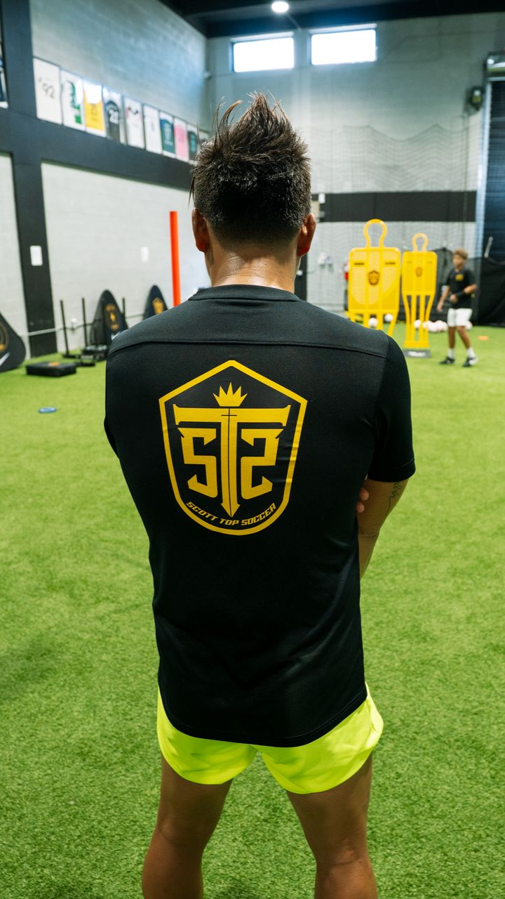 Person in black shirt with gold logo, standing on green turf. Yellow shorts. Gym setting.