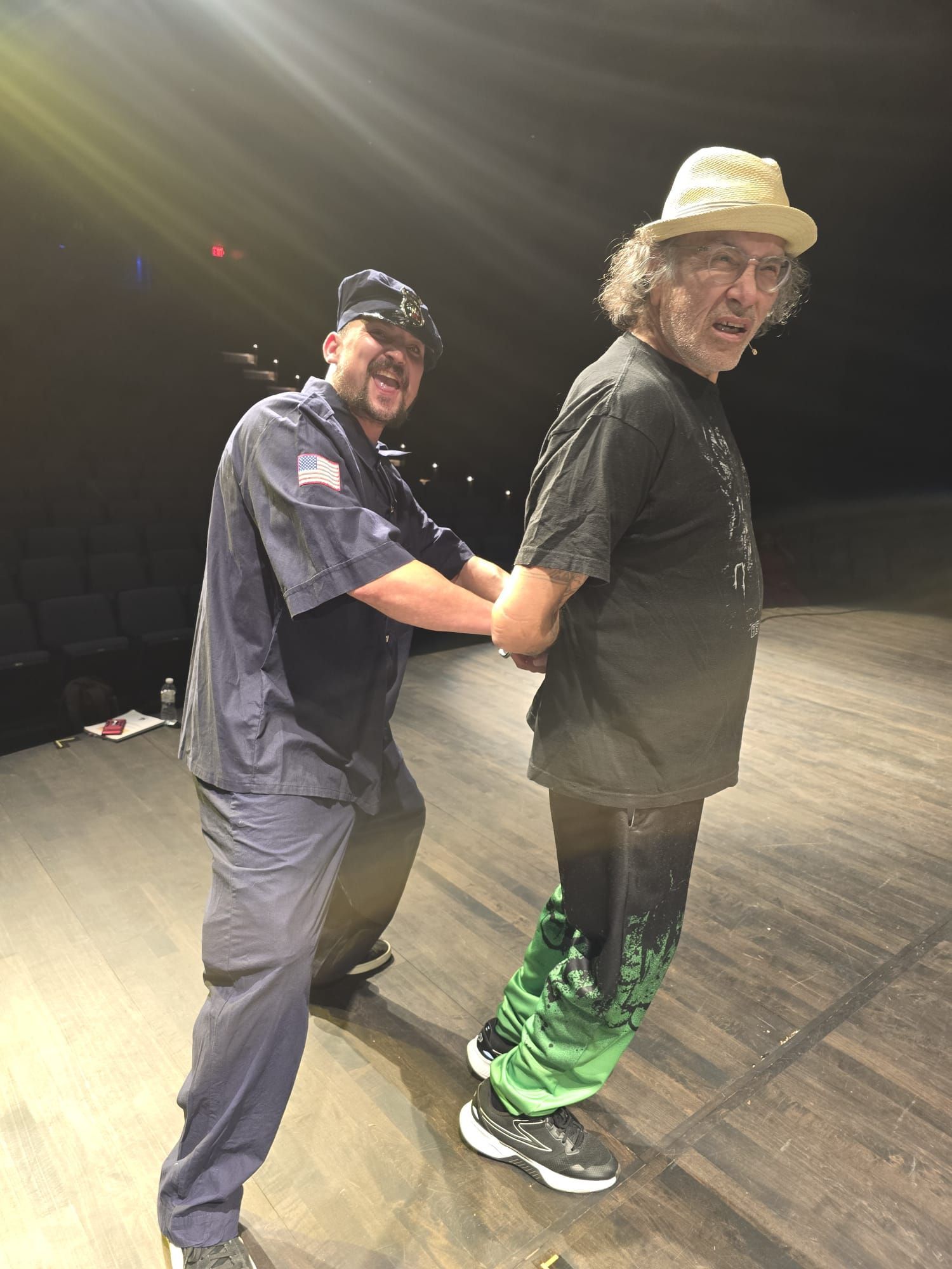 Two people on a stage, one playfully standing behind the other under bright overhead lights.