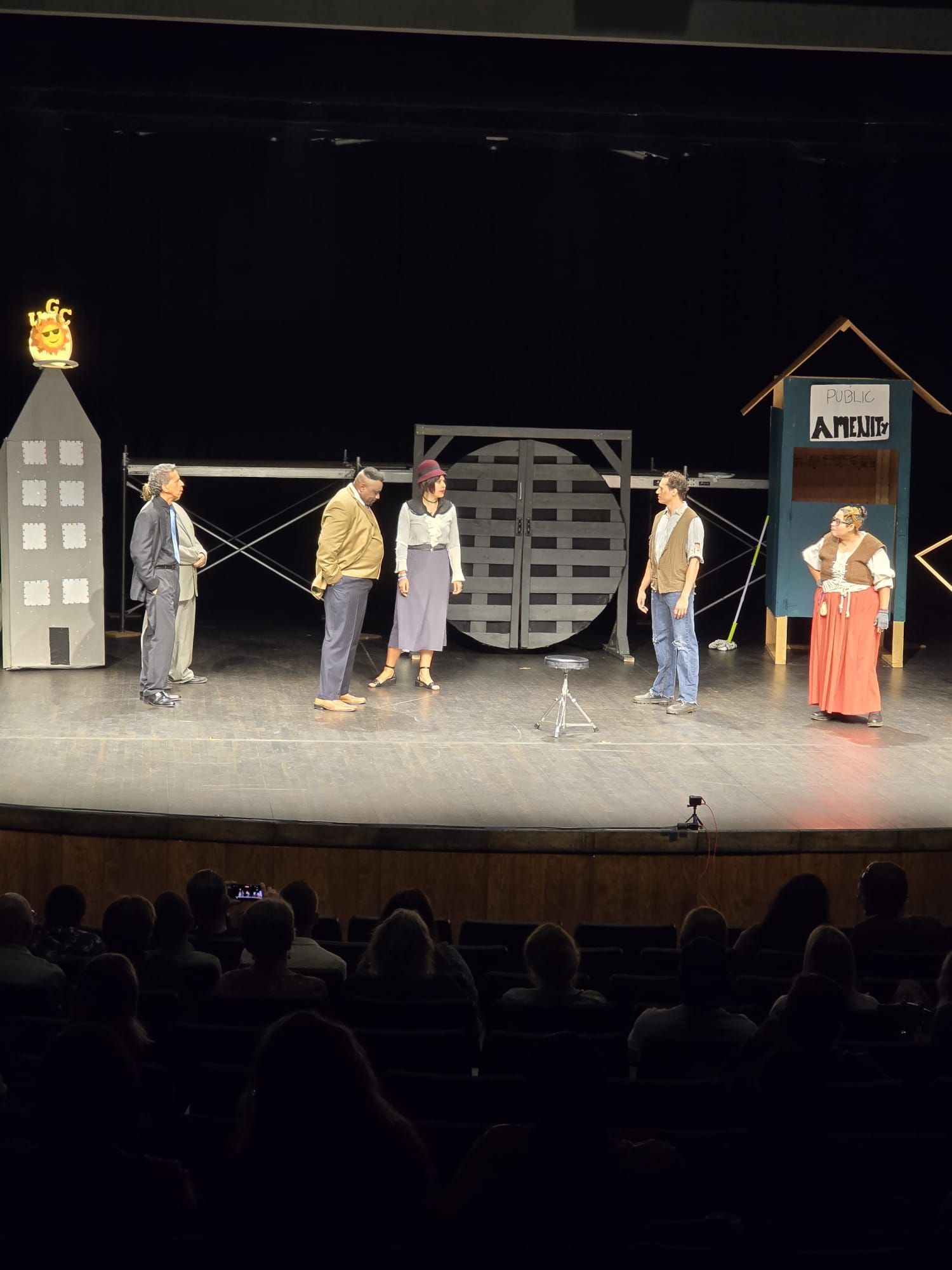 Five people stand on a theater stage with a cityscape set and an “APOL” sign, audience visible below.