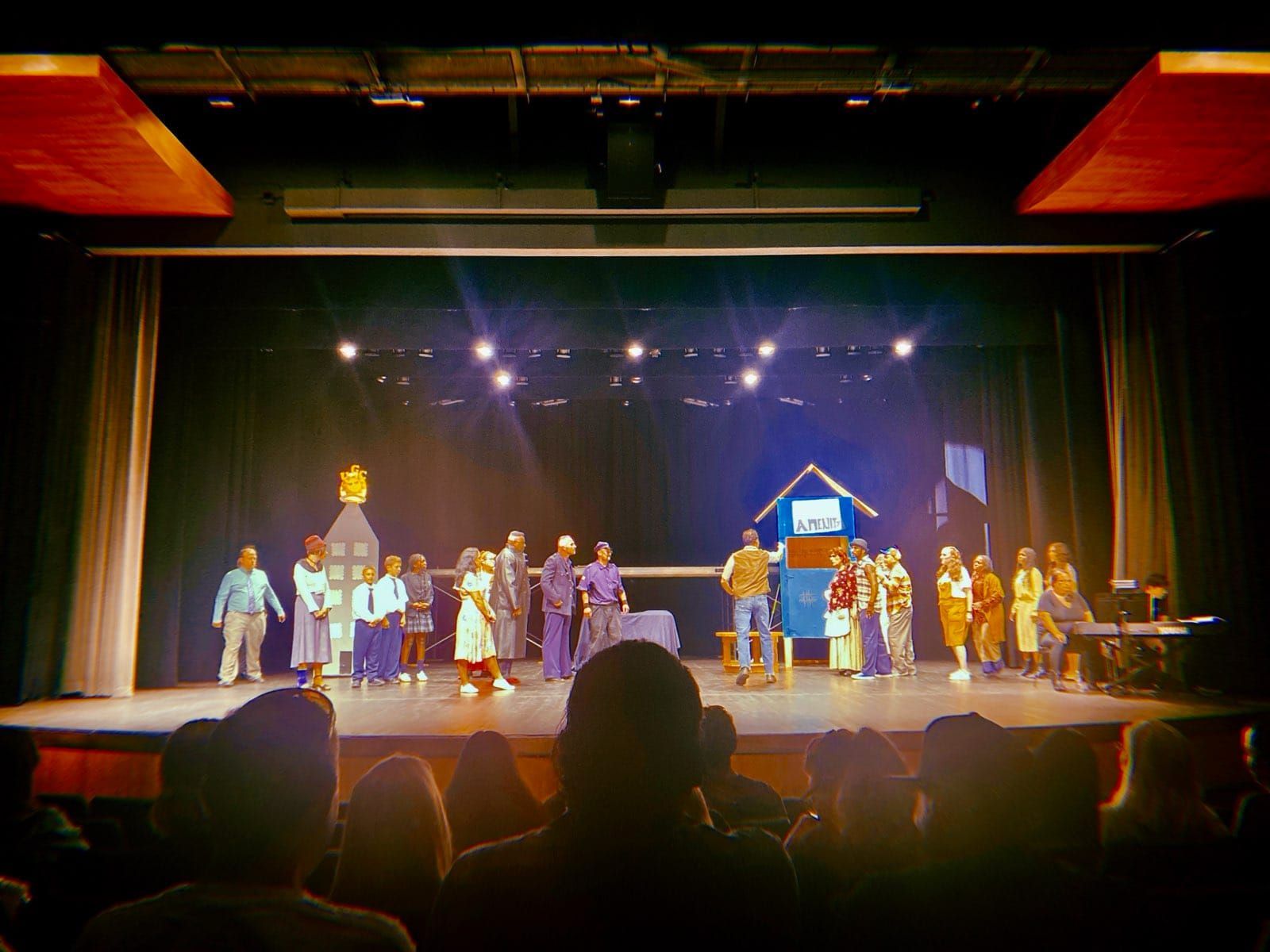 Performers on a theater stage under purple lights, with an audience silhouetted in the foreground.