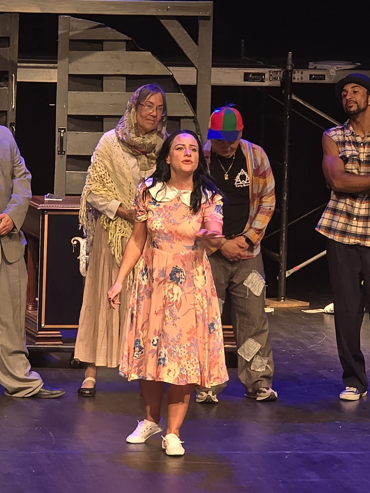 Actors on a stage set, with one woman in a floral dress standing front and center.