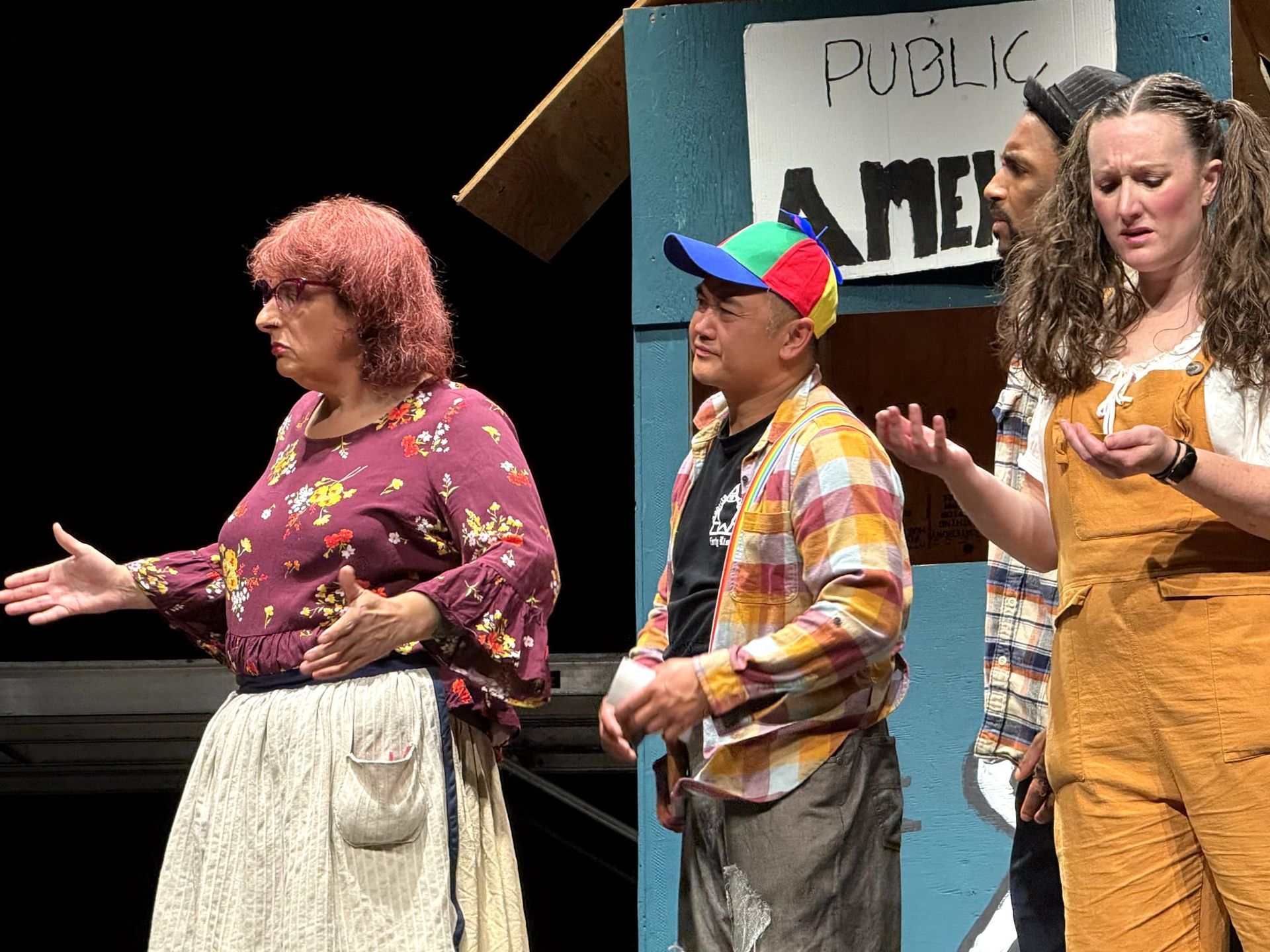 Three performers on stage, including a person in a multicolored cap and two in orange overalls, gesturing during a show