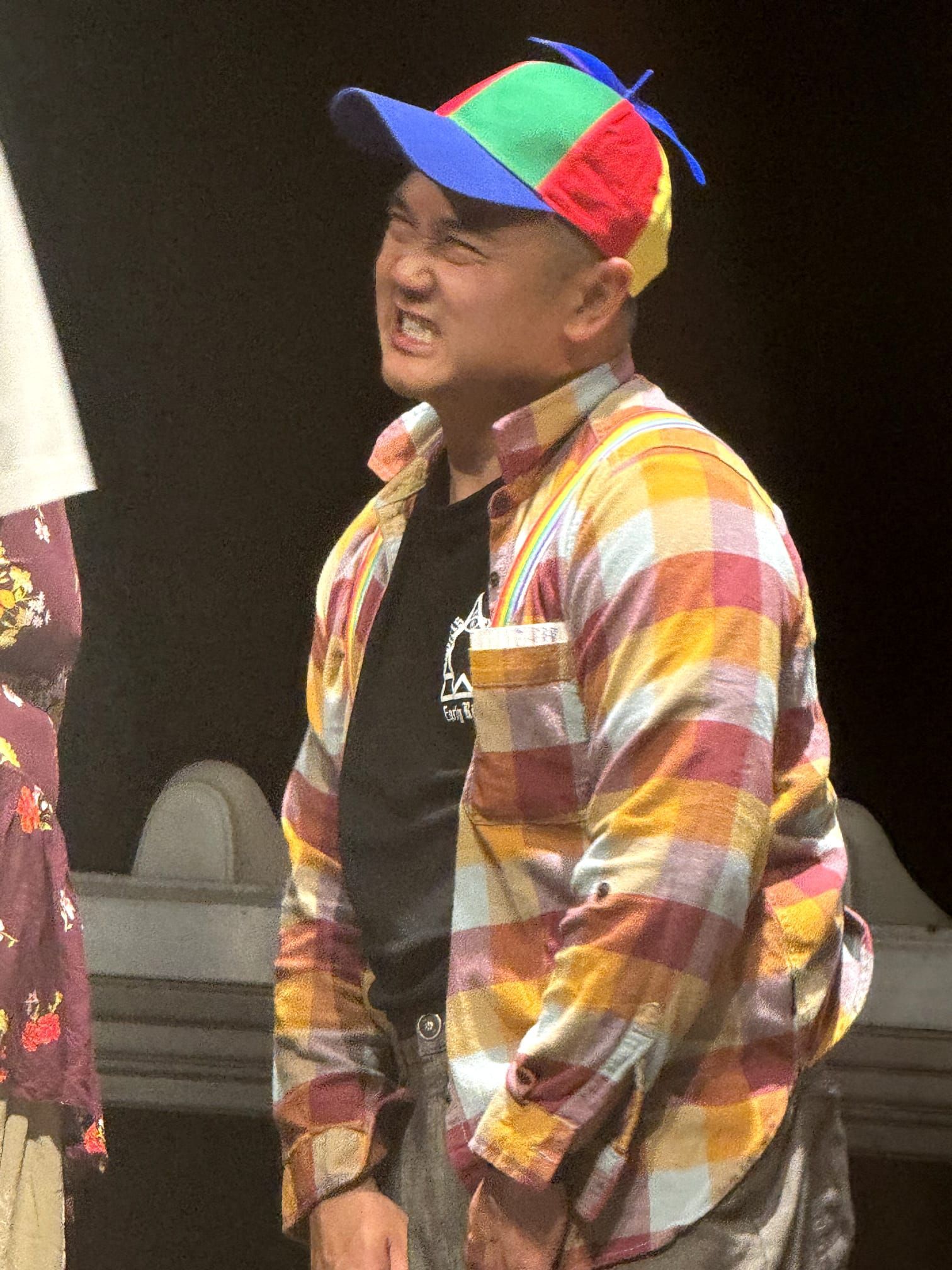 Person in a multicolored cap and plaid shirt smiling on stage, hands in pockets.
