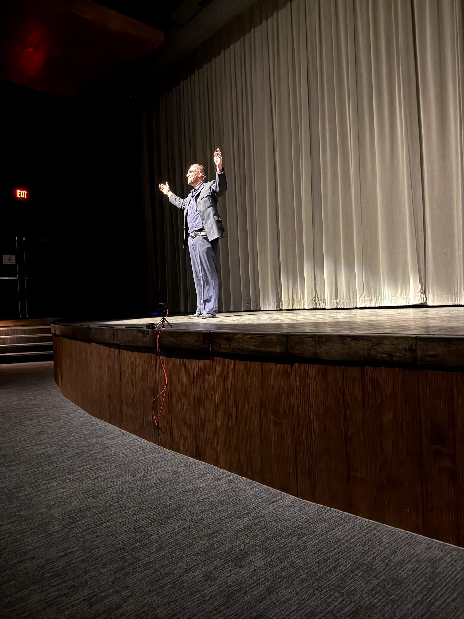 Person standing on a theater stage beside a closed curtain, raising both arms under spotlights.