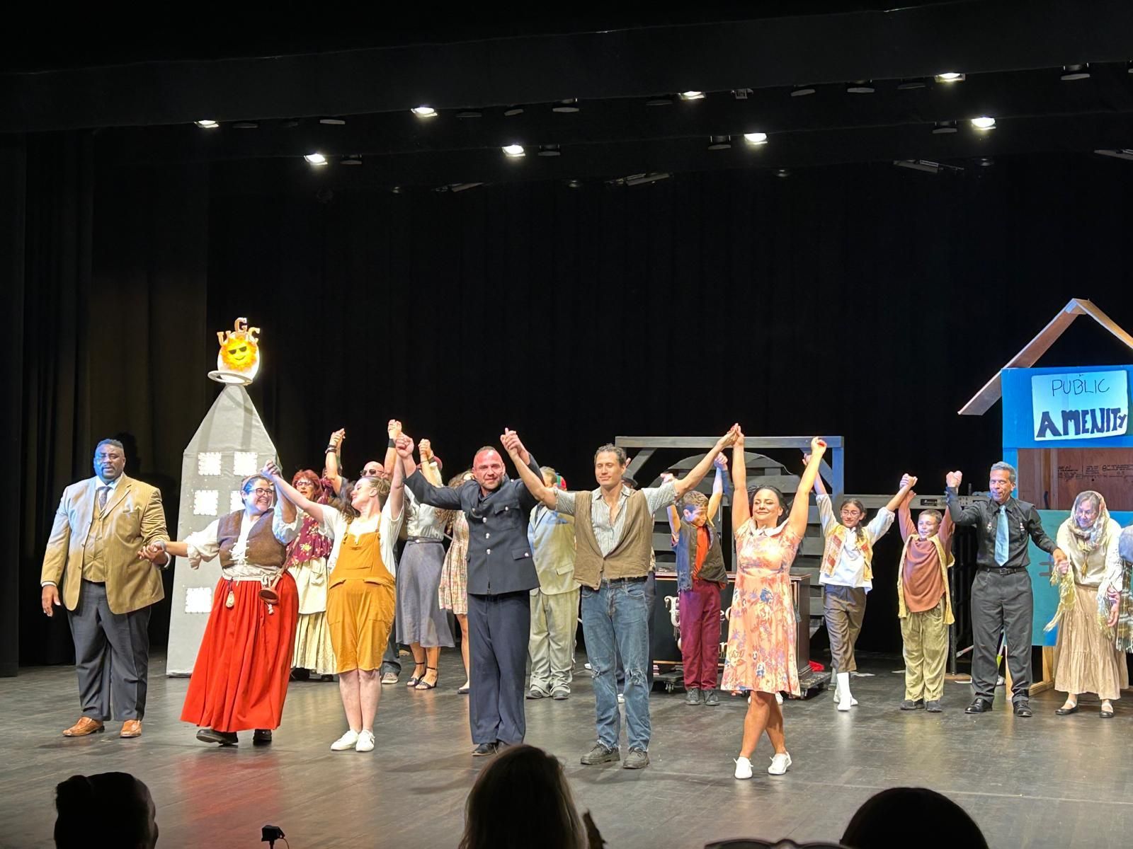 Group of performers onstage holding hands with arms raised under bright lights, with a house prop and sign in back