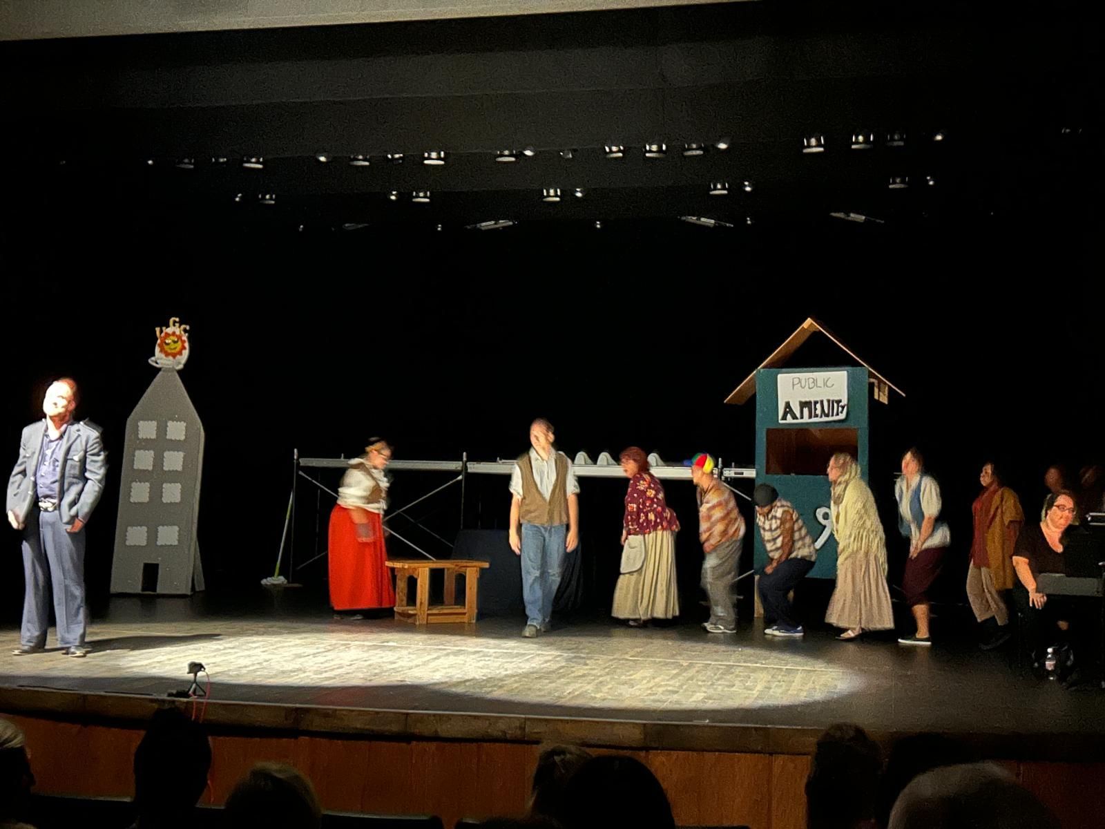 Theater stage with a line of costumed actors, a house prop, and a tall cutout building on a dark backdrop