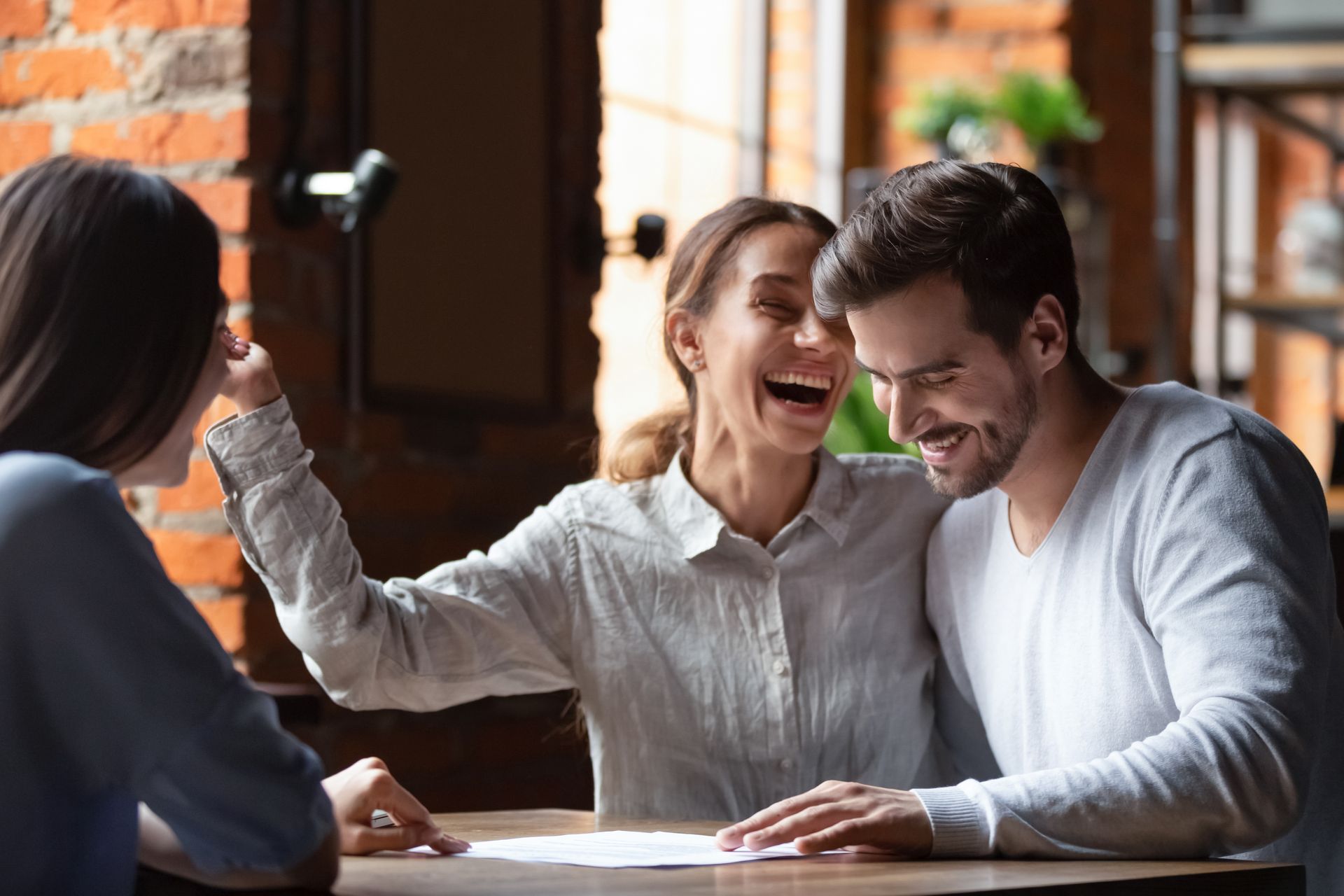 Three coworkers laughing at a café table, one raising a fist while looking at a laptop