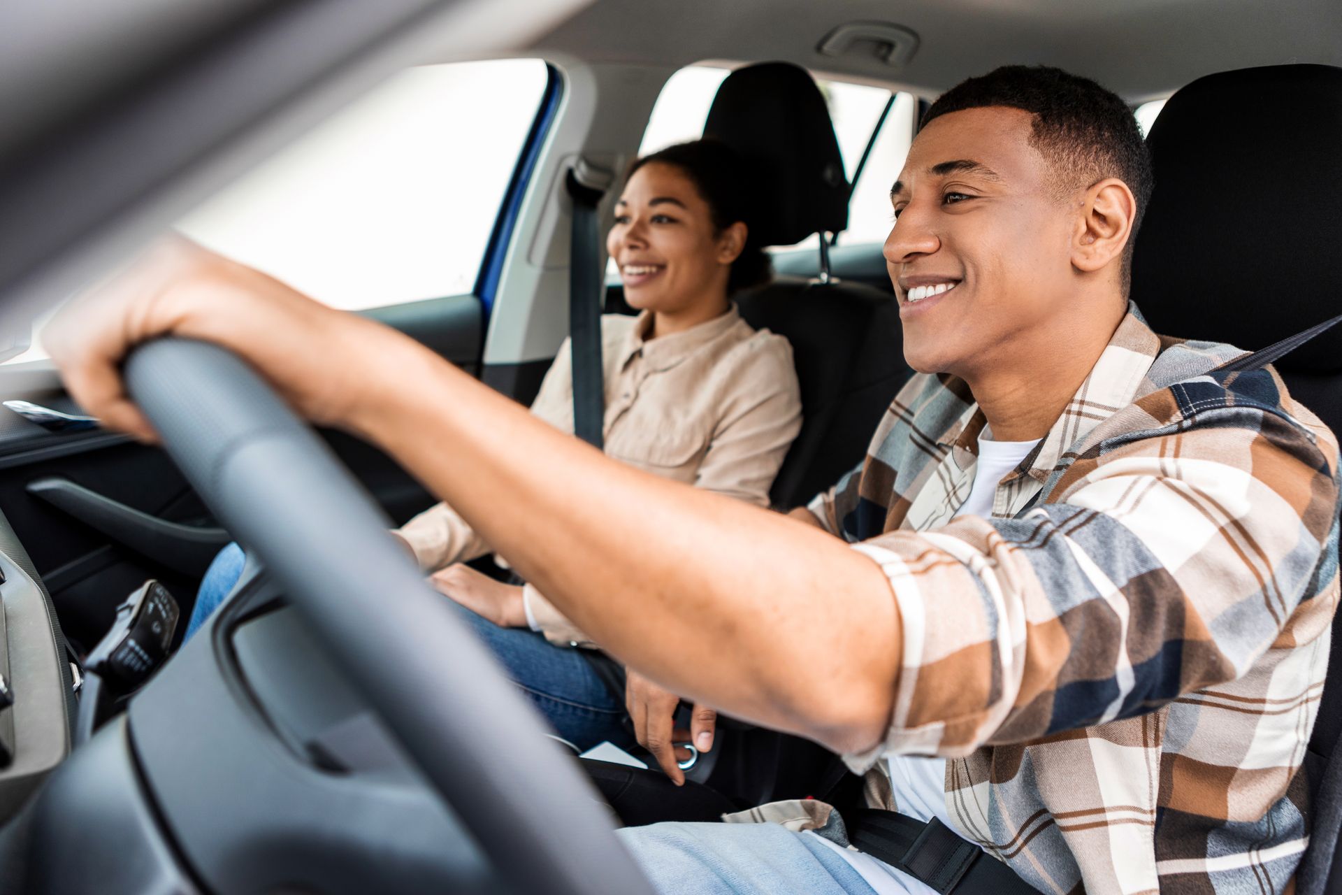 Two smiling people riding in a car, with the driver holding the wheel and the passenger seat visible.