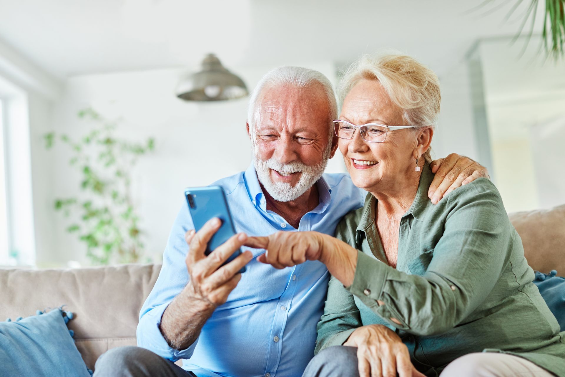 Smiling elderly couple on a couch looking at a smartphone in a bright living room