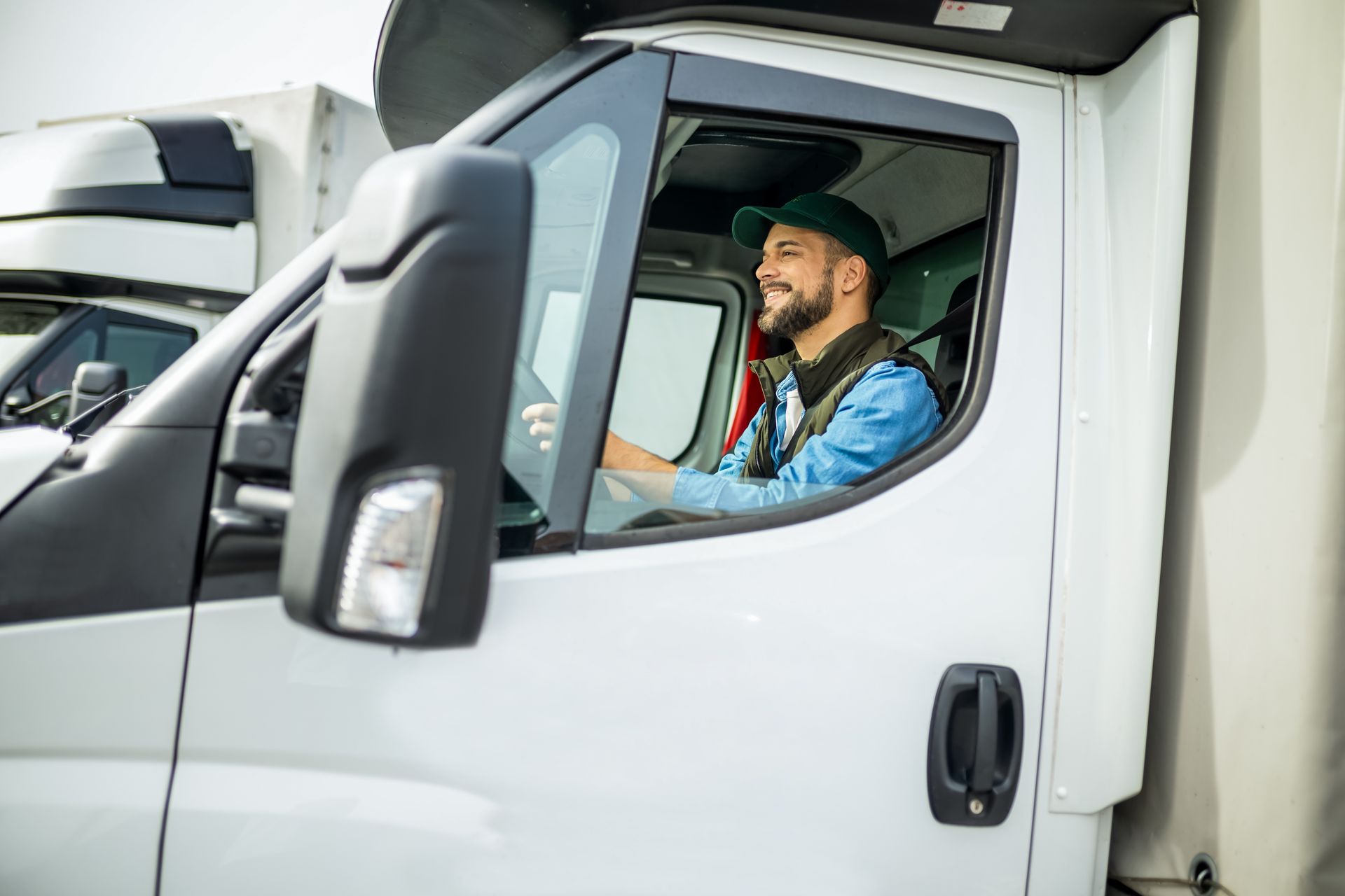 Truck driver smiling in a white cab, wearing a blue jacket and red vest.