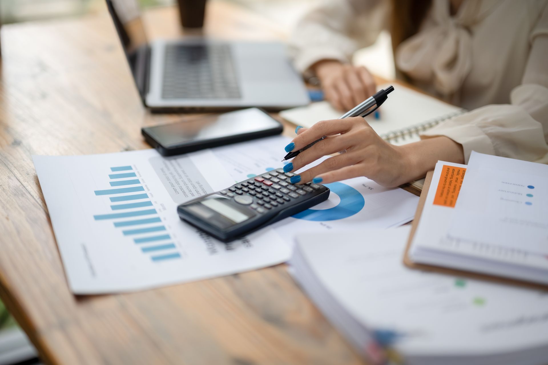 Person using calculator at a desk with charts, laptop, phone, and paperwork