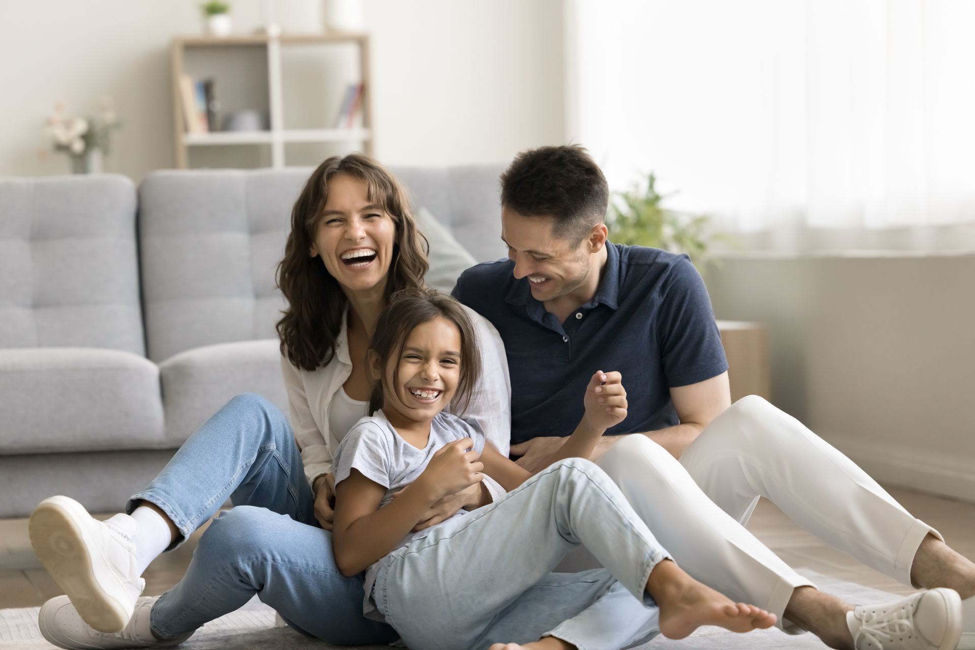 Family laughing together on a living room floor beside a gray sofa
