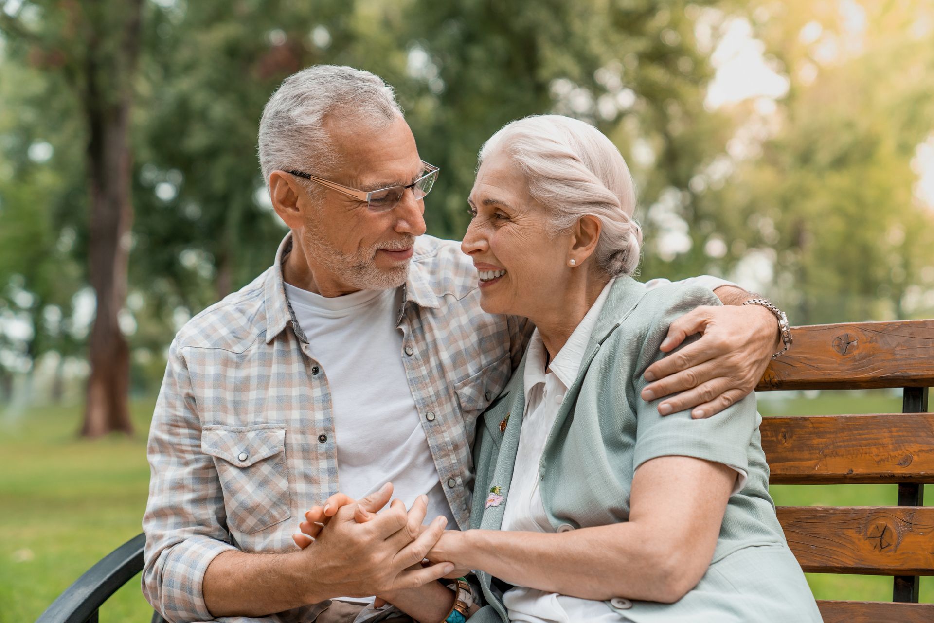 Two elderly people sit on a park bench, smiling and holding hands in a green outdoor setting.