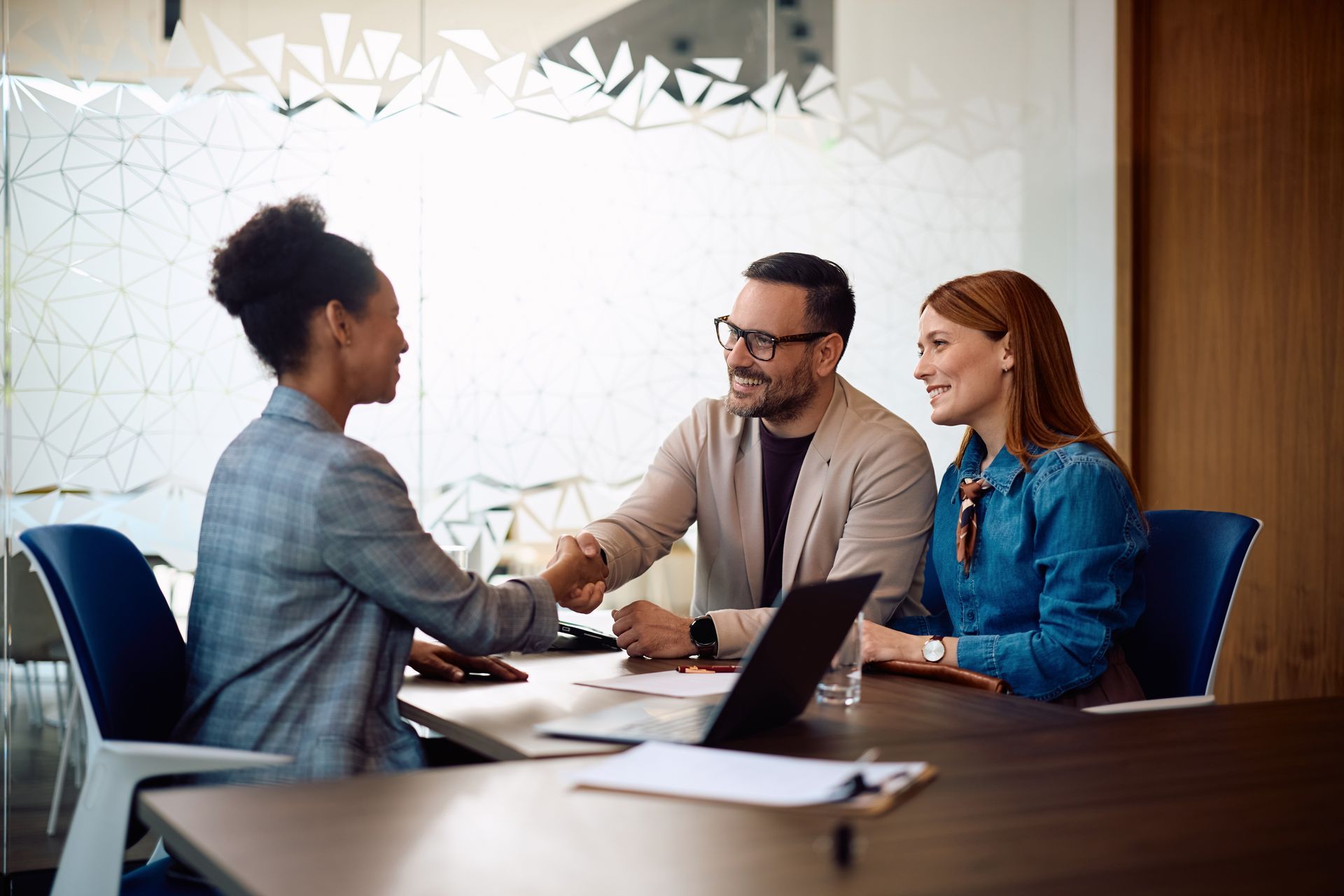 Three coworkers in a meeting room shaking hands across a table with a laptop and papers