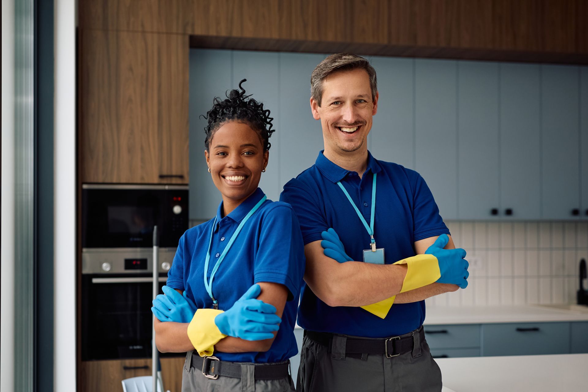 Two smiling cleaners in blue uniforms with yellow gloves stand in a modern kitchen.