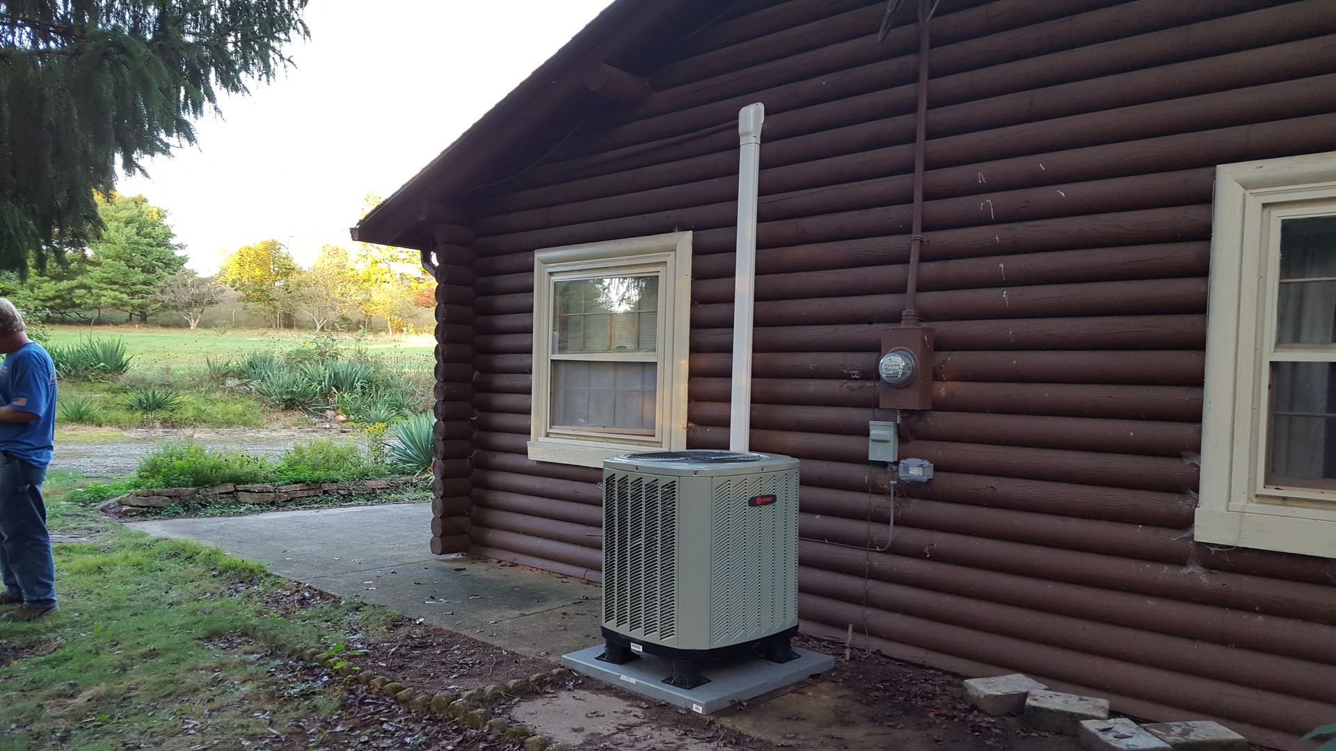 Outdoor HVAC unit beside a brown log-cabin wall, with two windows and a person standing at left.