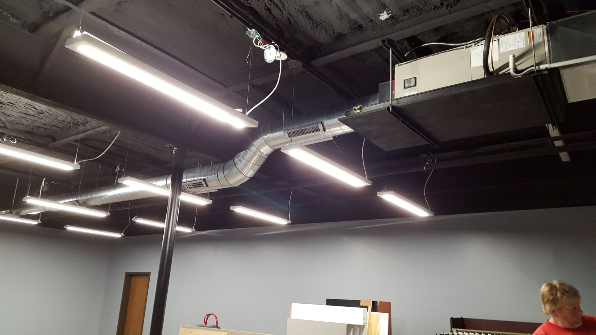 Office ceiling with exposed ducts and bright strip lights; two people are partially visible below.