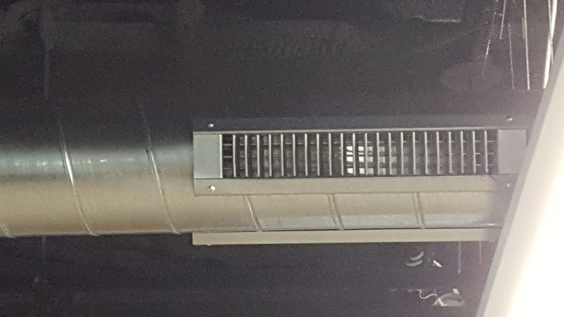 Close-up of a metallic air duct with a rectangular vent grille in a dim ceiling space