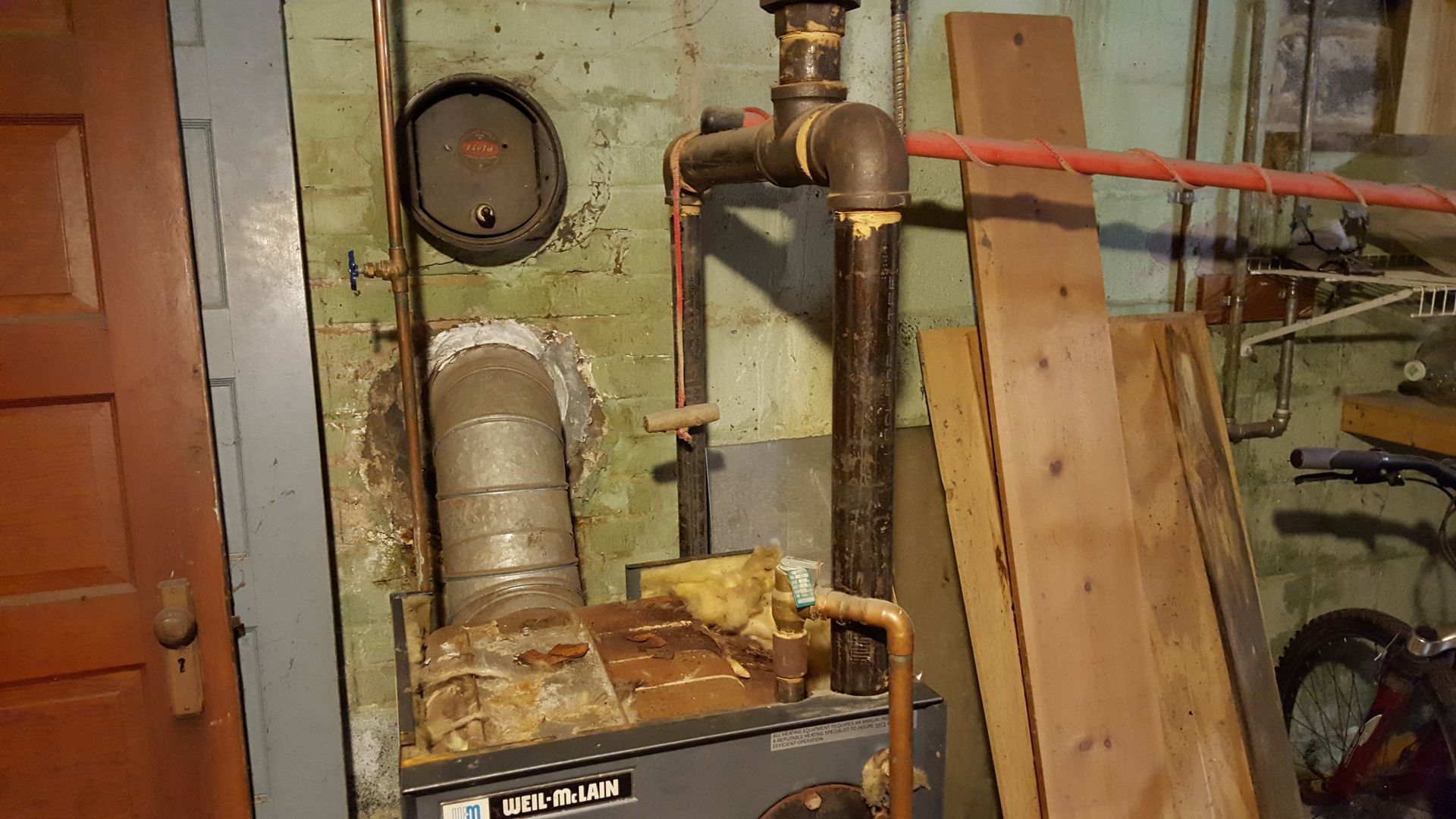 Basement utility area with old furnace, exposed pipes, and leaning boards against a brick wall