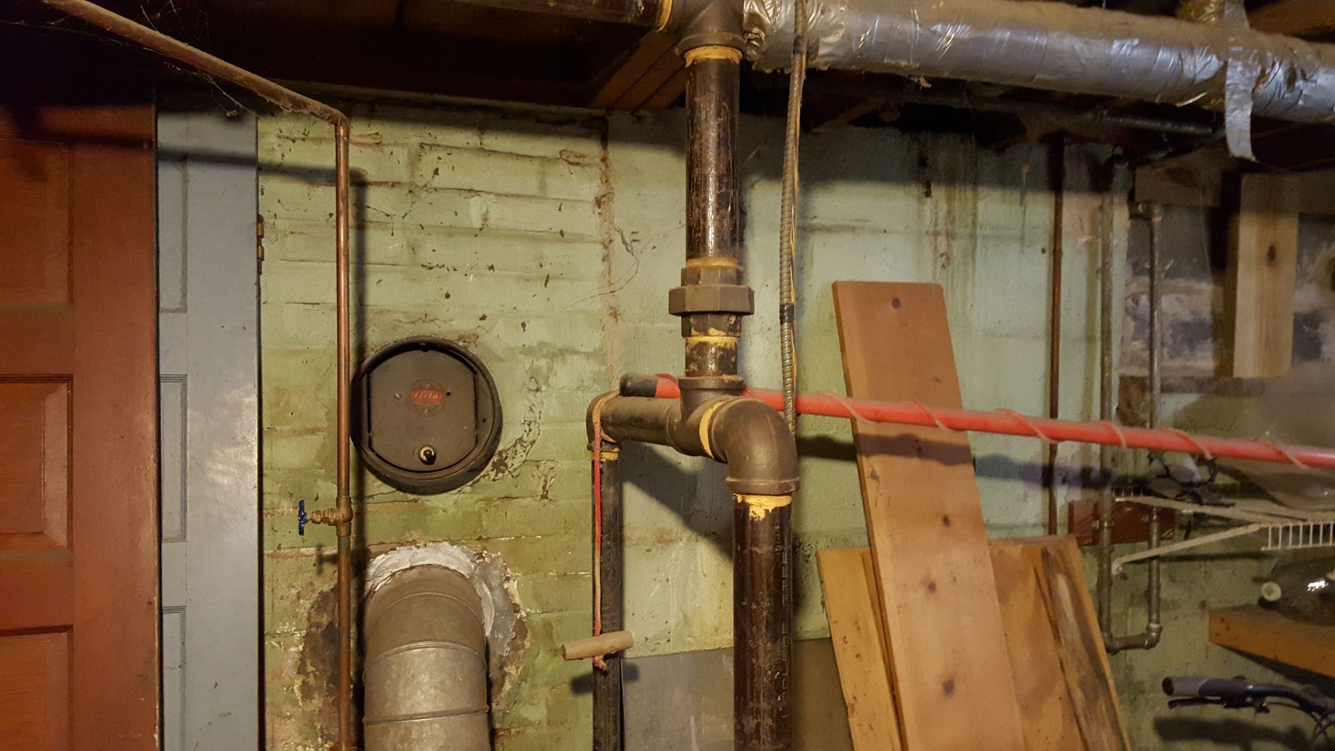 Basement utility room with exposed pipes, valves, a round meter, and wooden boards leaning against the wall.