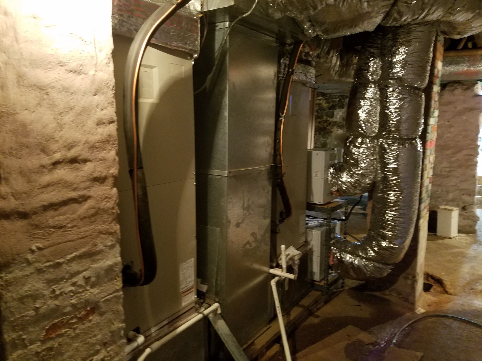Basement utility room with insulated ductwork, metal pipes, and a large HVAC unit along rough stone walls.