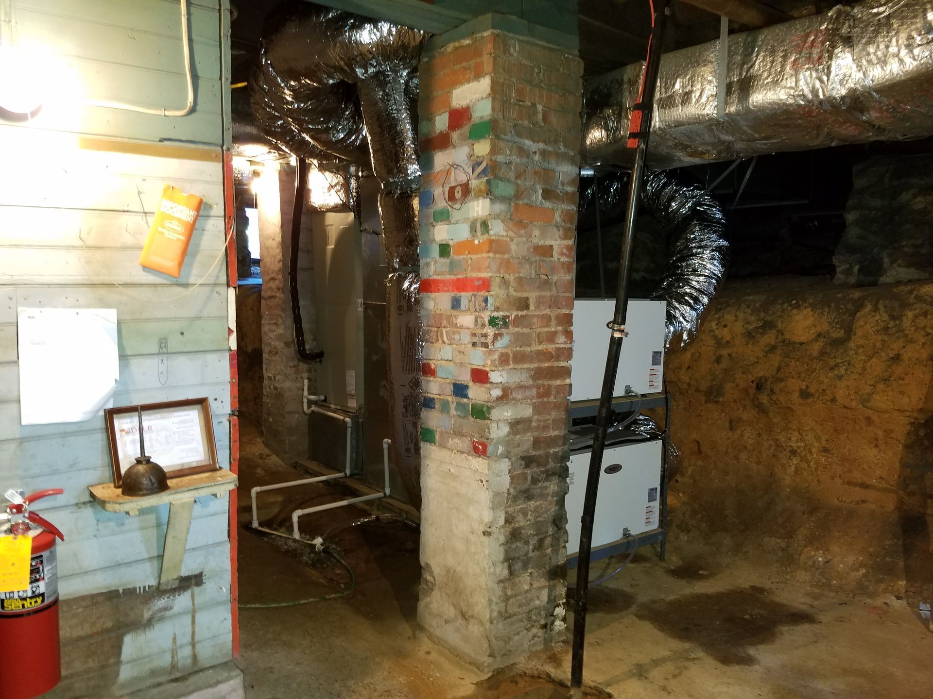 Dim basement with exposed pipes, brick column, utility boxes, and a small work area.