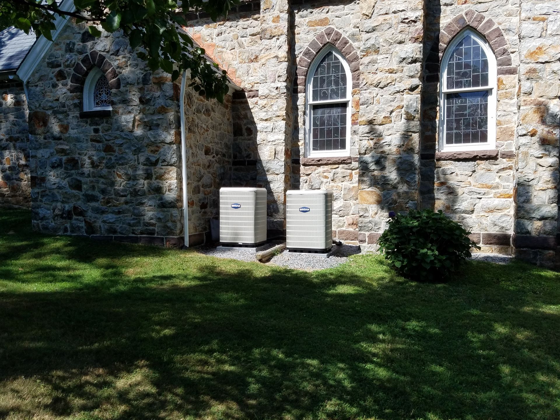 Stone church wall with two white utility boxes on a shaded grassy lawn, framed by arched windows.