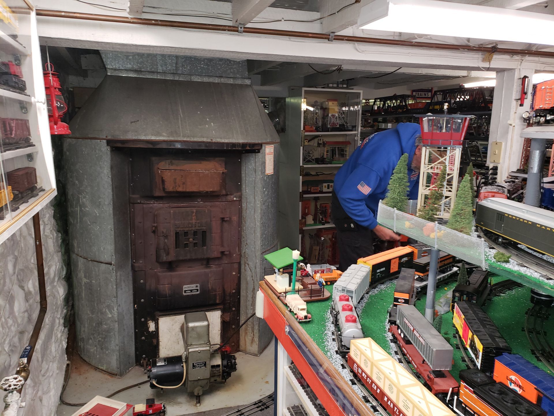 Person in blue jacket inside a cluttered miniature train workshop with model tracks and buildings