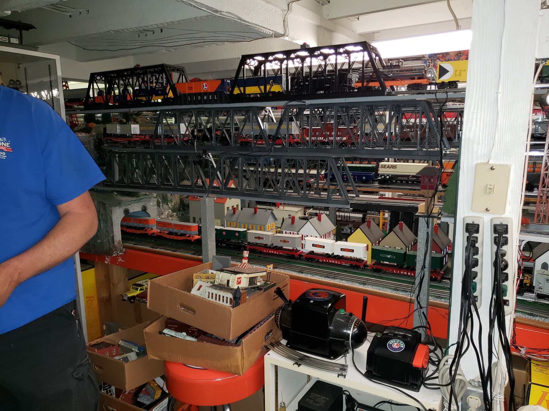 Shelves packed with electronic equipment and cables in a workshop, with boxes and tools on the floor.